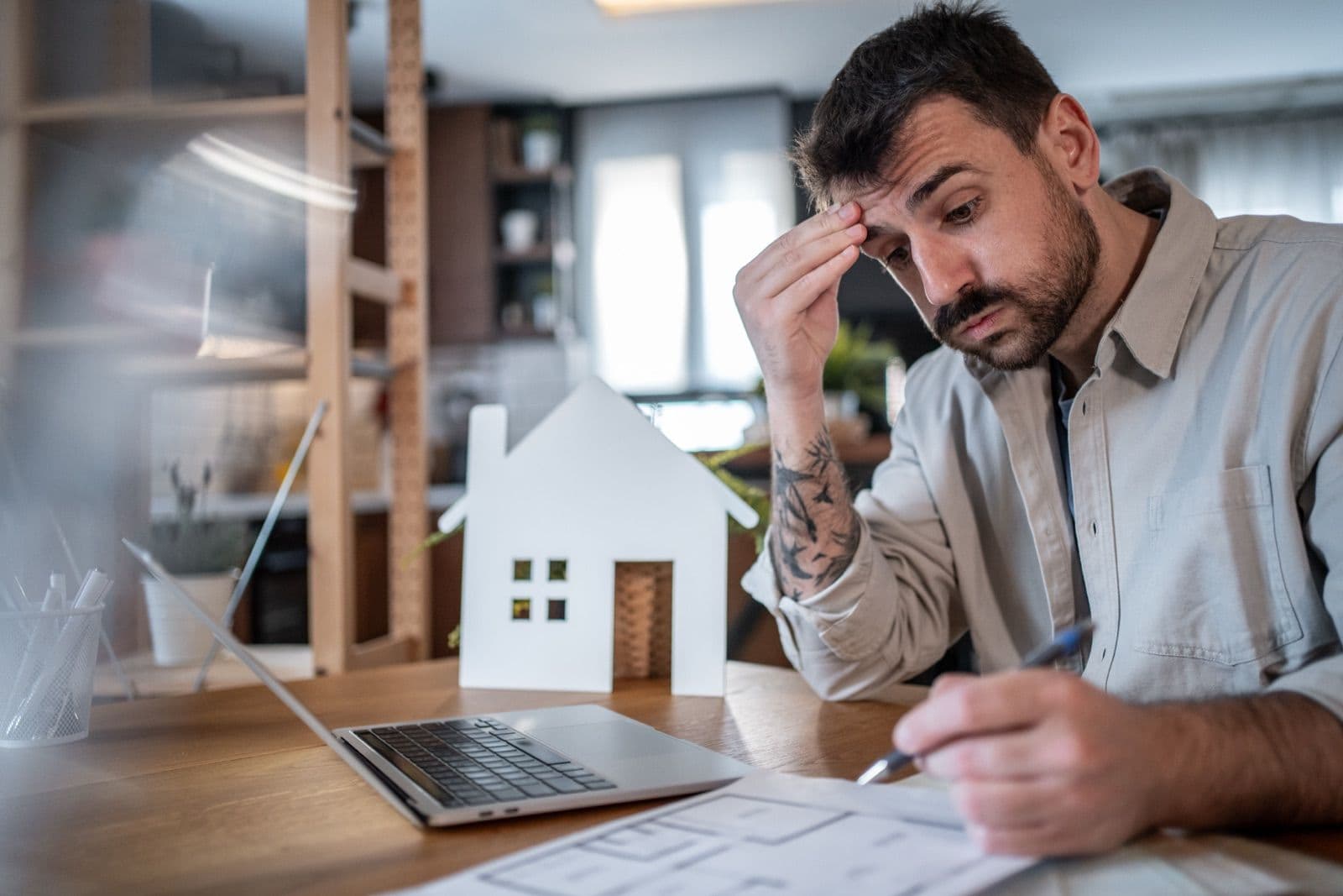 Man at home looking stressed over paperwork, a small model house on his desk