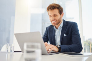 One happy young caucasian businessman working on a laptop in an office. Confident male entrepreneur planning while browsing the internet and sending emails to clients. Man joining a virtual conference via webcam