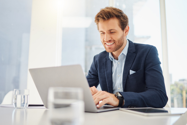 One happy young caucasian businessman working on a laptop in an office. Confident male entrepreneur planning while browsing the internet and sending emails to clients. Man joining a virtual conference via webcam